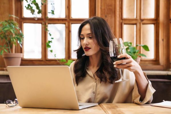 Image of serious adult woman drinking red wine and working with laptop while sitting in cozy cafe indoors